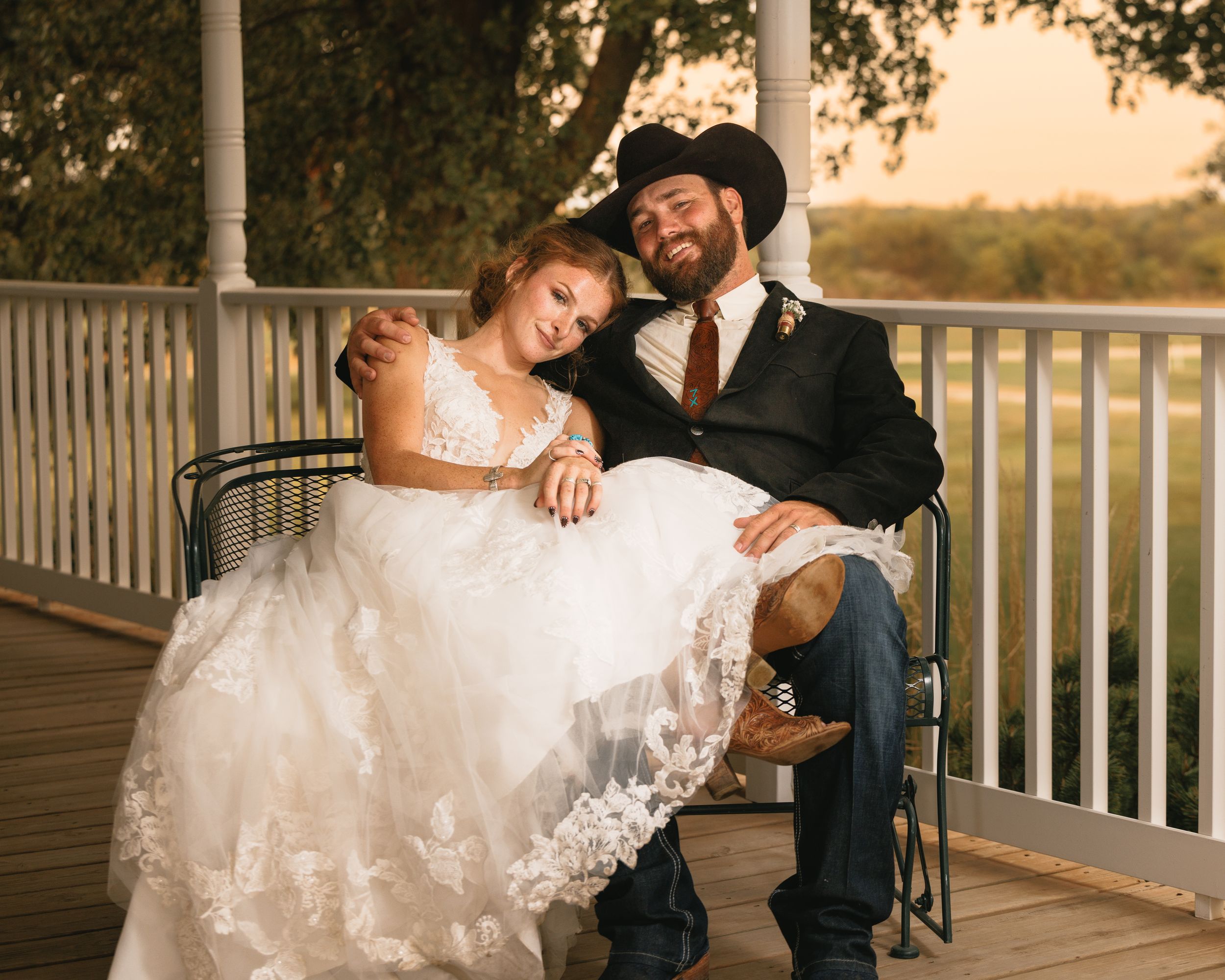 Bride in lace wedding gown leans on groom in black cowboy hat on rustic porch at golden hour.