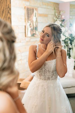 A bride wearing a white lace wedding dress puts on pearl earrings while getting ready for her special day.