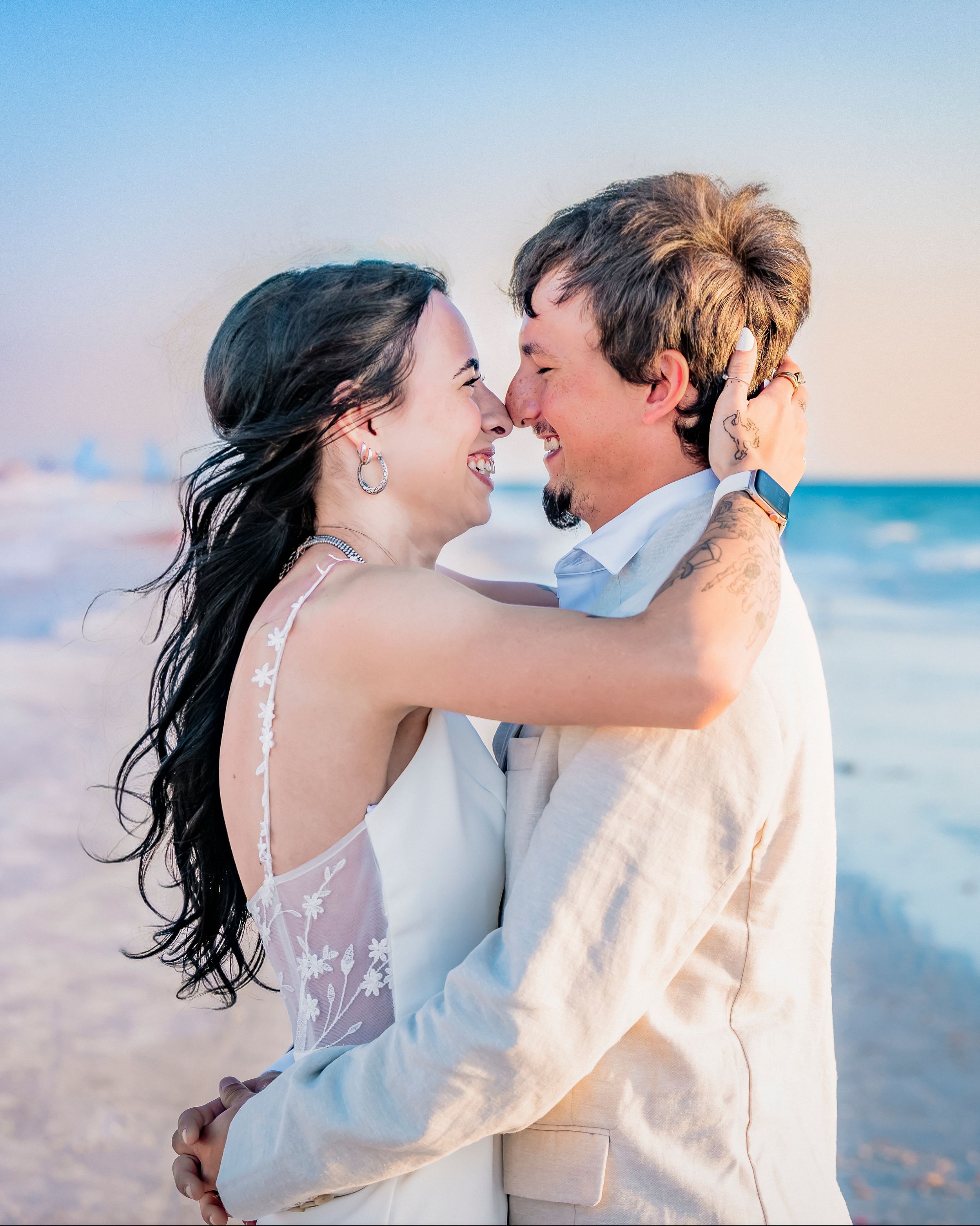 A joyful couple embraces on a beach at sunset, the bride in a white lace gown and groom in a tan suit. Florida beach destination wedding