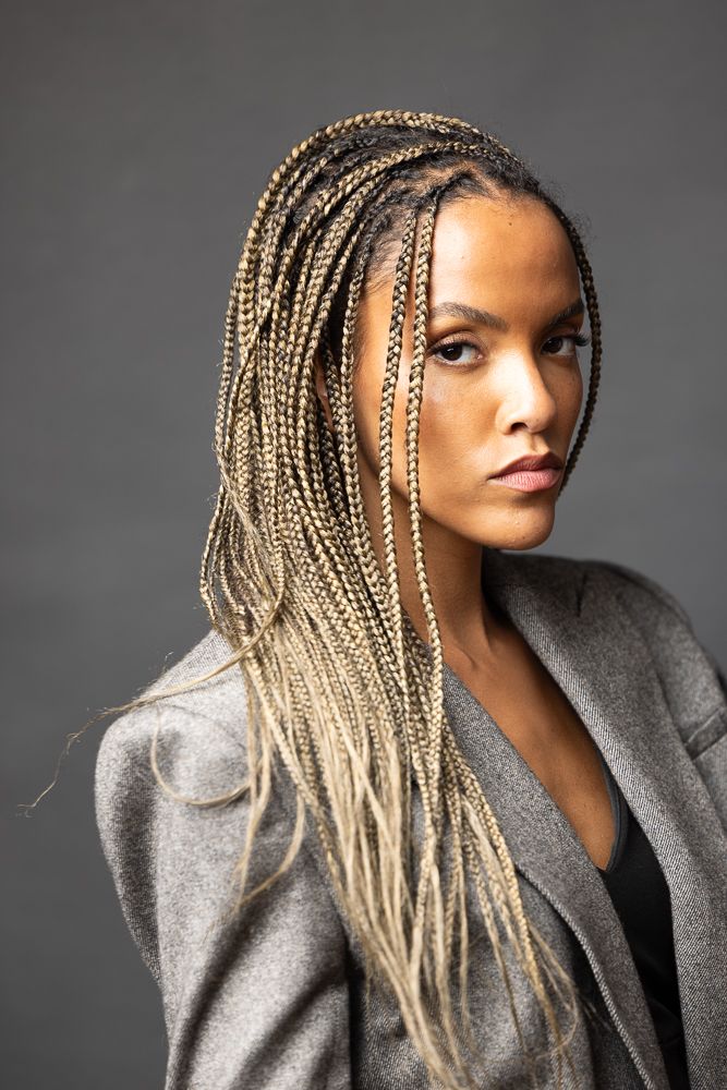 Young woman with long blonde box braids wearing a gray blazer, posing against a dark gray background.