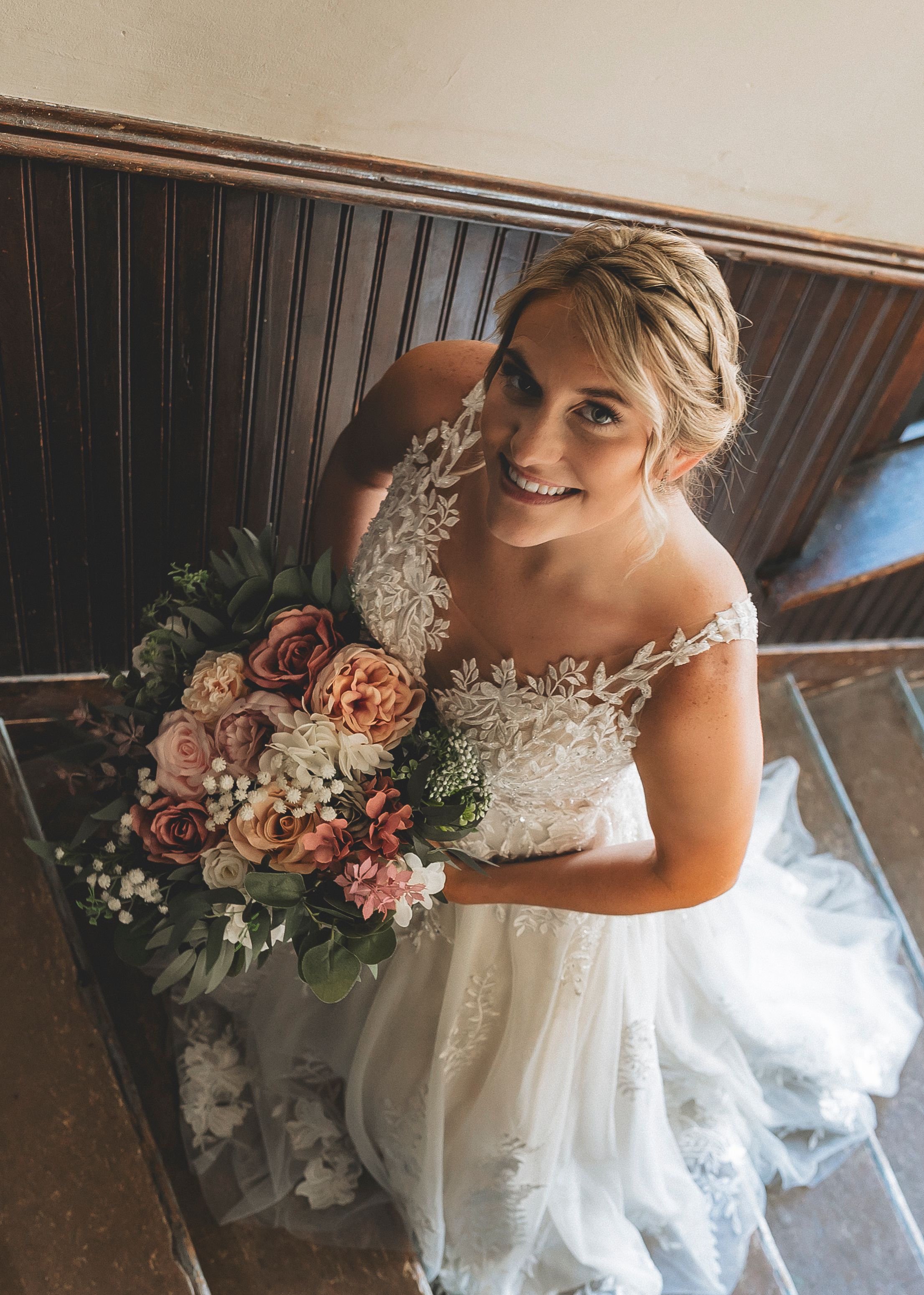 A bride in a lace wedding dress carries a romantic bouquet of pink and peach roses with white flowers and greenery.