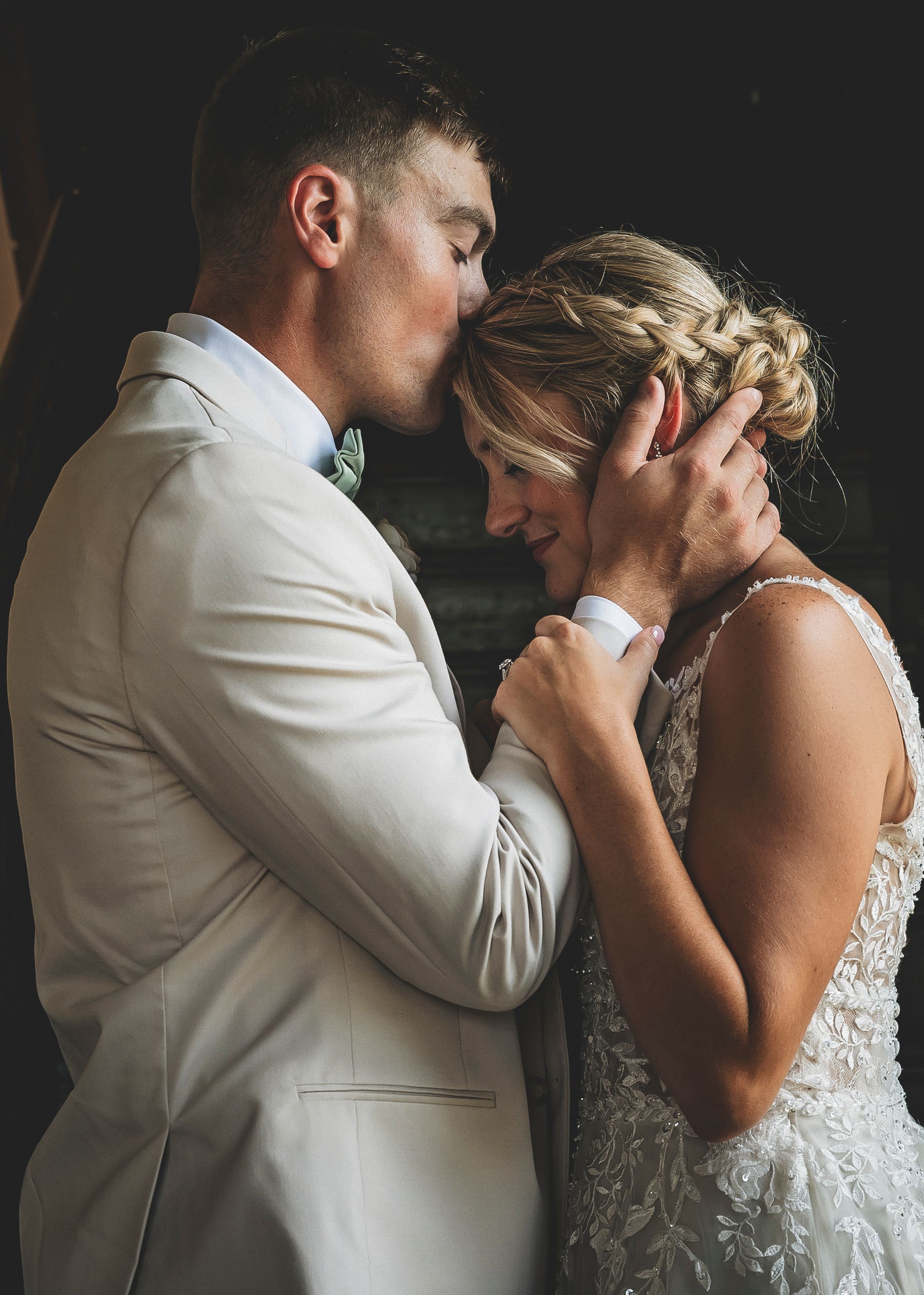 A tender romantic moment between newlyweds in wedding attire sharing an intimate embrace against a dark background.