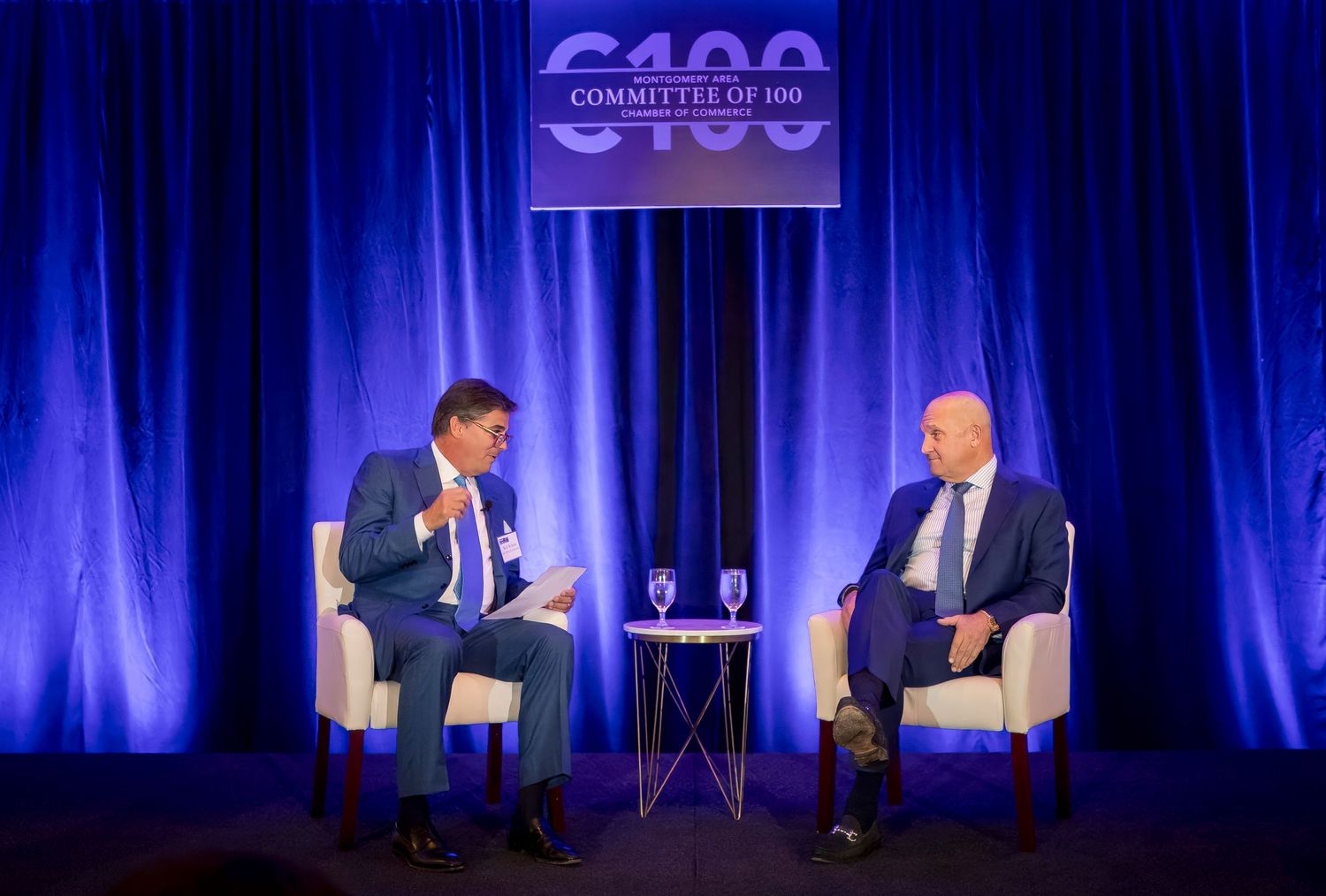 Two speakers on stage at C100 event with blue lighting and white chairs.