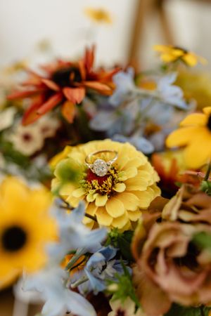 A diamond engagement ring nestled in a yellow zinnia flower with other blooms in soft focus.