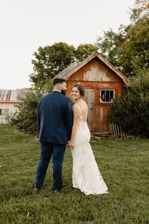 Wedding couple poses on grassy field with rustic barn in background during golden hour.