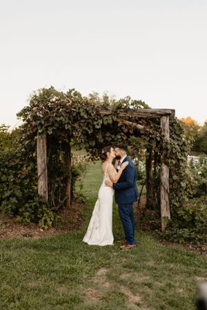 A romantic wedding couple embraces under a rustic wooden arch covered in greenery and vines.