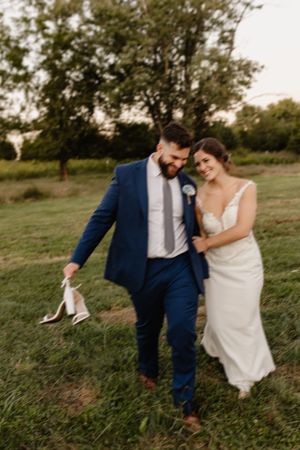 A couple walking together on grassy field during sunset with the bride holding her shoes.