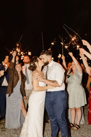 Wedding couple sharing a kiss surrounded by guests holding sparklers during nighttime celebration.