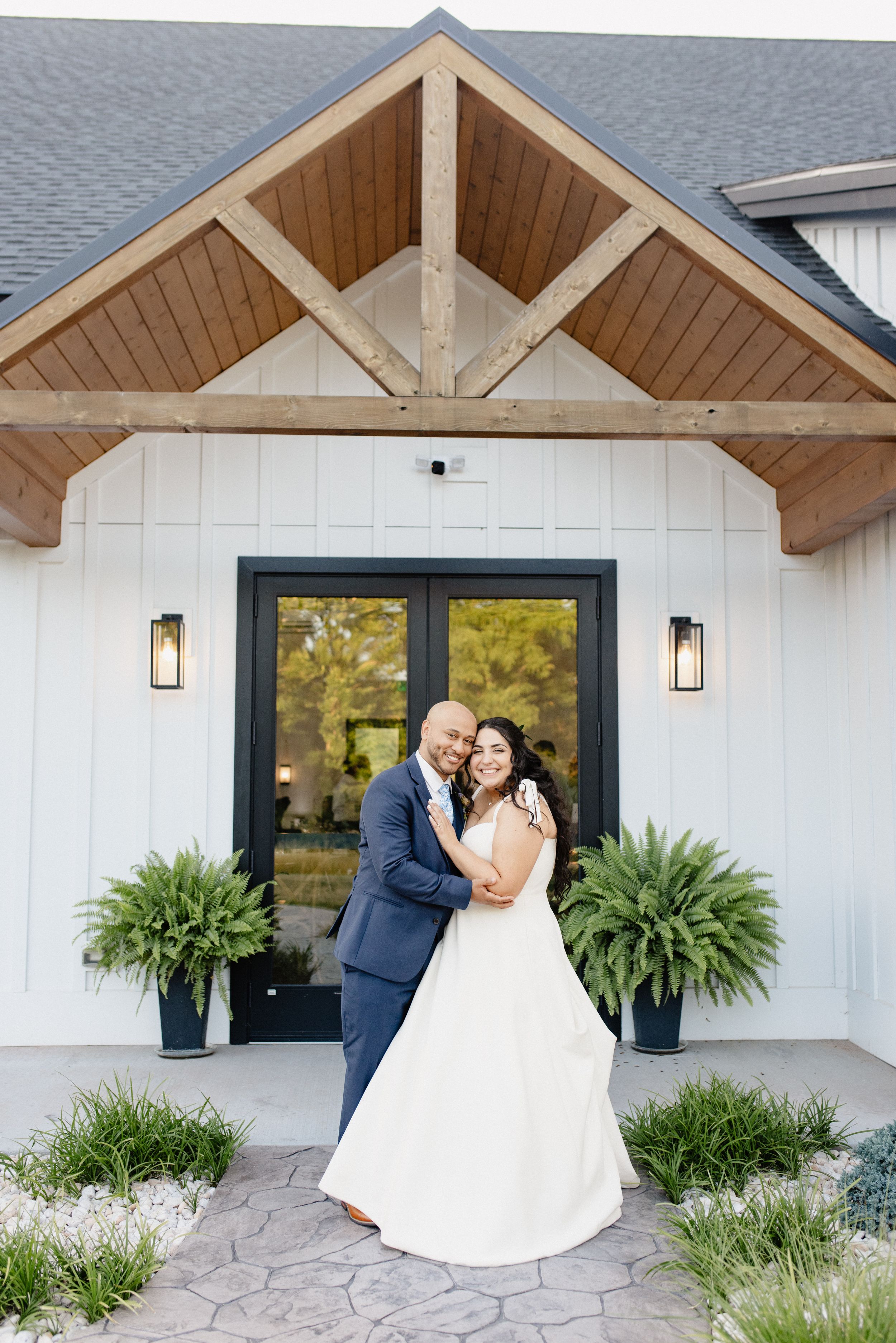 A modern farmhouse entrance with architectural details and potted ferns frames a couple.