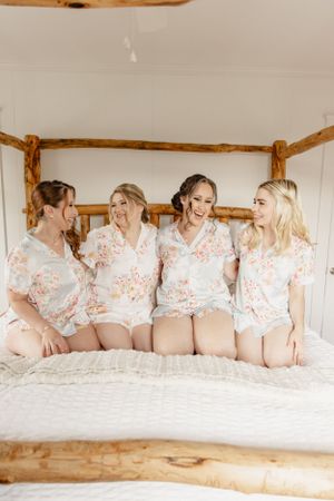Bridesmaids in floral robes pose playfully on bed during wedding preparation.