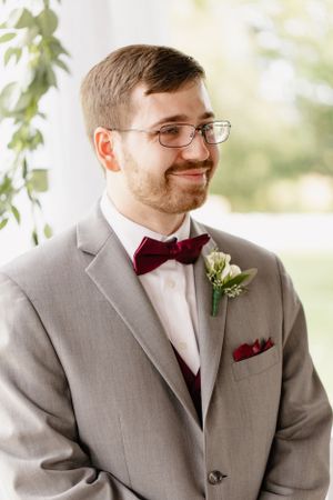 Groom in grey suit with burgundy bowtie smiles in natural light setting.