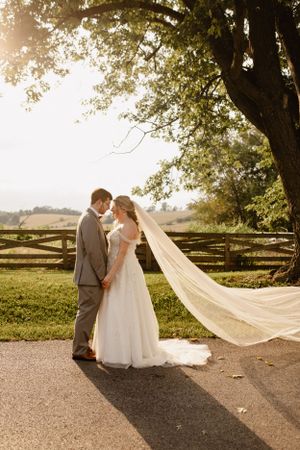 Wedding couple poses together outdoors with bride's veil flowing in sunlit countryside setting.