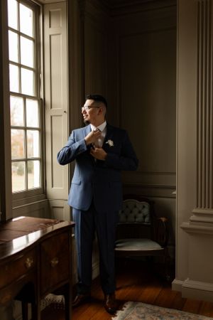 A groom in a navy blue suit adjusts his tie while standing by a window in an elegant room.