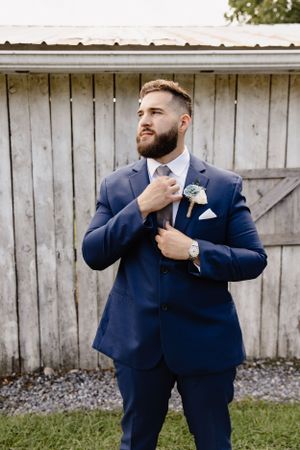 A groom in navy blue suit adjusts his tie while standing against a rustic white barn.