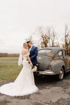 A wedding couple poses beside a vintage grey car on a cloudy autumn day.