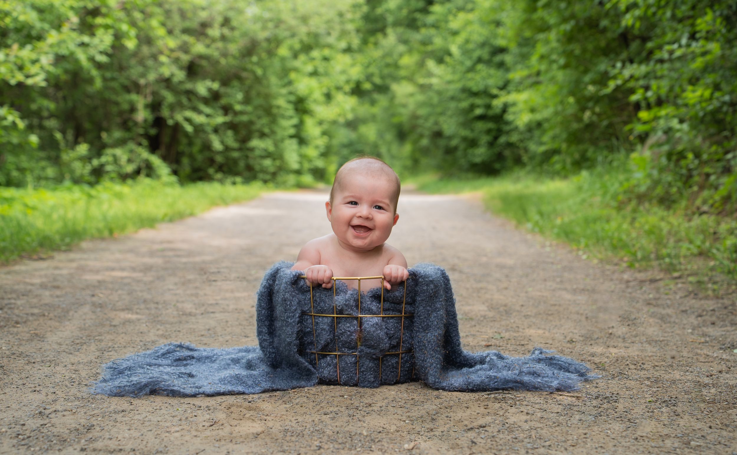 Small baby sitting in denim overalls on a dirt path surrounded by green foliage.