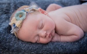 Peaceful sleeping newborn rests on soft grey blanket wearing delicate blue floral headband.