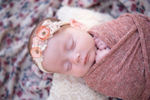 Sleeping newborn swaddled in dusty rose wrap wearing peach floral headband on floral print backdrop.