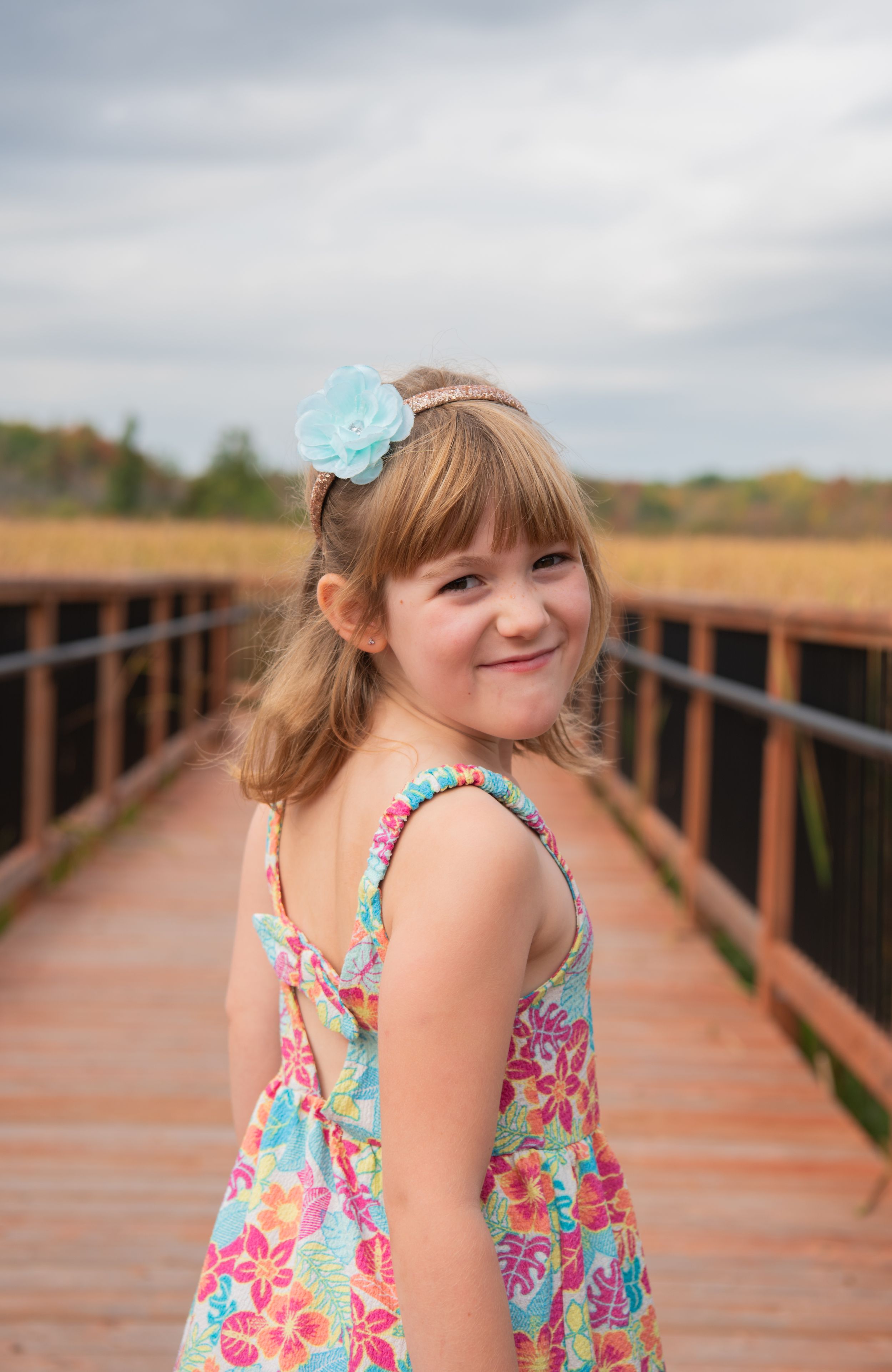 A young child in a floral dress smiles while standing on a wooden boardwalk on a cloudy day.