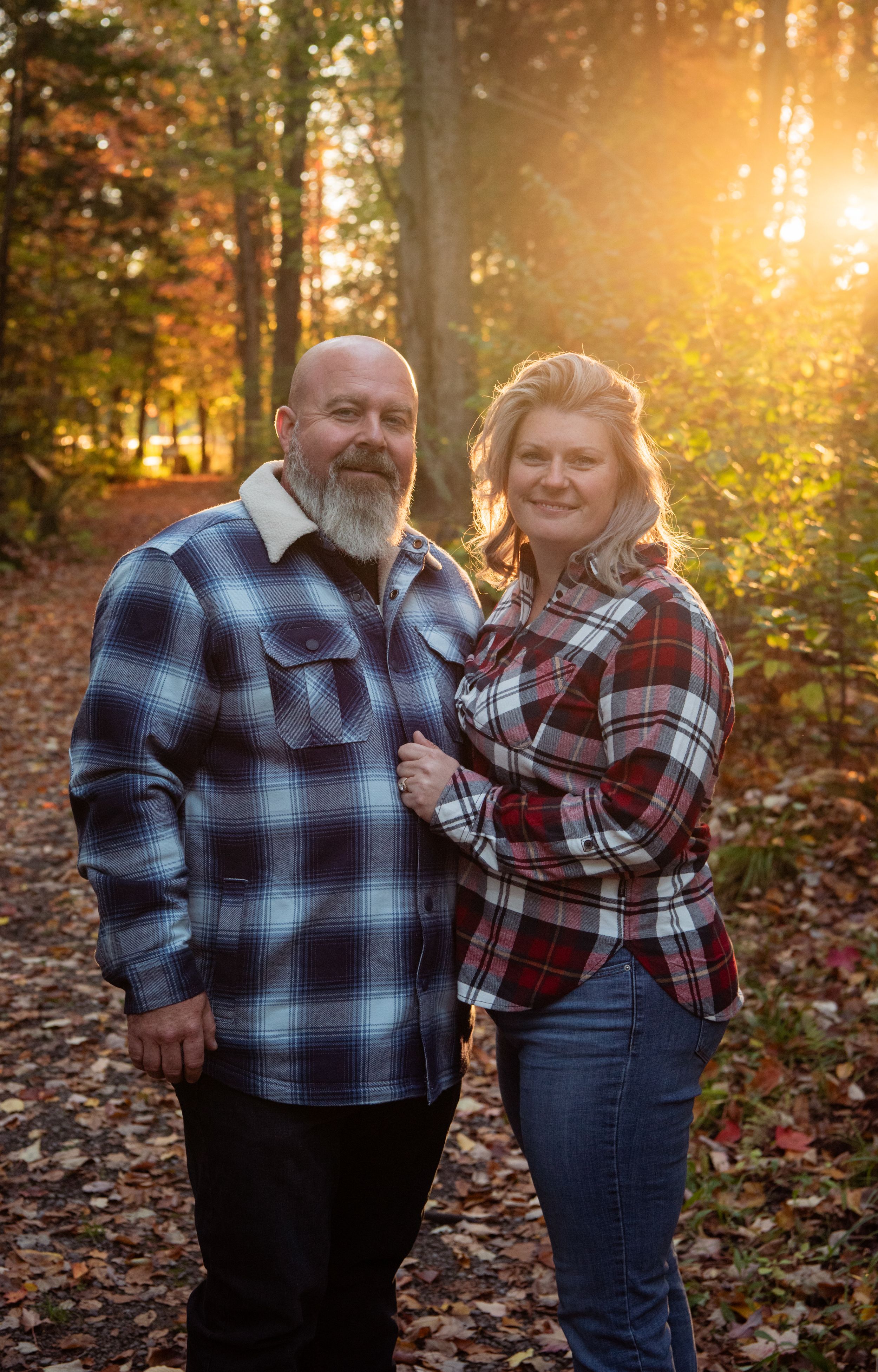 A couple wearing matching plaid flannel shirts pose together in a sunlit autumn forest with fallen leaves on the ground.