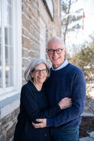 A mature couple embraces and smiles outside their stone house in winter.