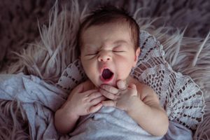 A newborn baby yawns while lying on lace and furry textured bedding.