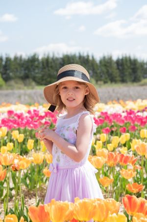 A child in a sun hat and lavender dress poses among orange and pink tulips.