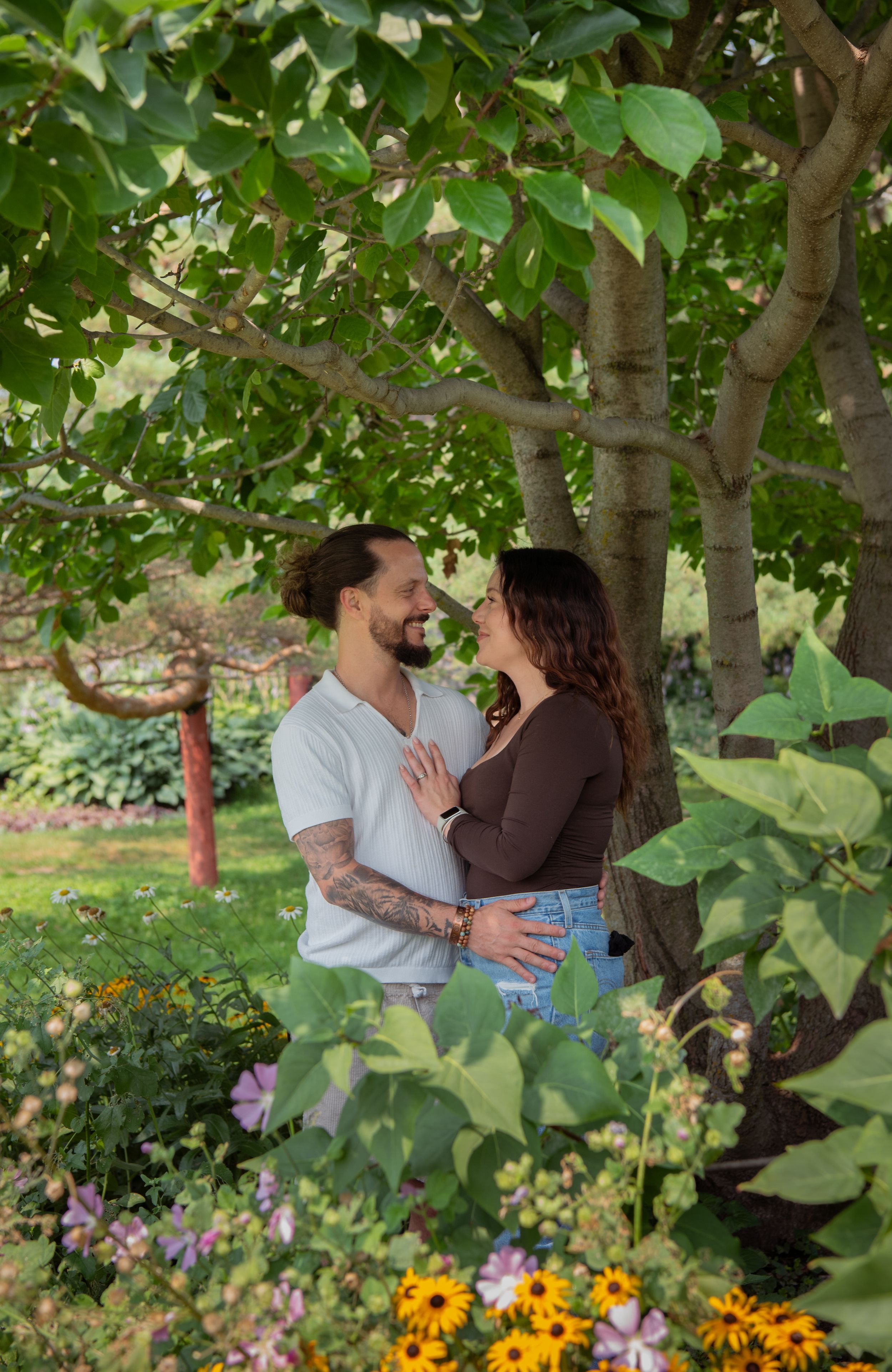 A couple shares a romantic moment in a garden surrounded by vibrant flowers and lush green trees.