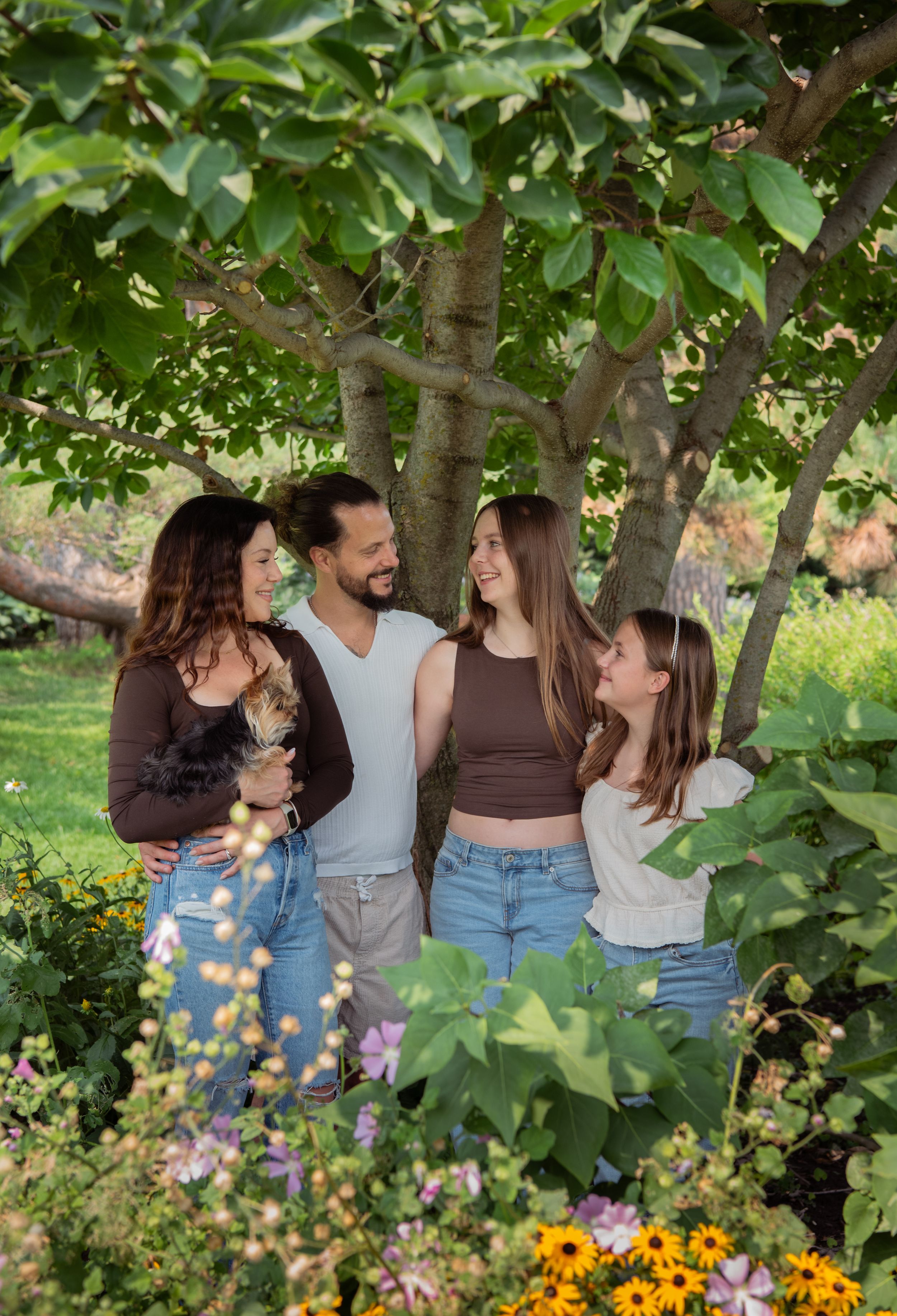 Three people share intimate moments in a lush garden setting with vibrant flowers and greenery.