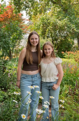 Two people stand together in a garden with blooming daisies and autumn foliage in the background.