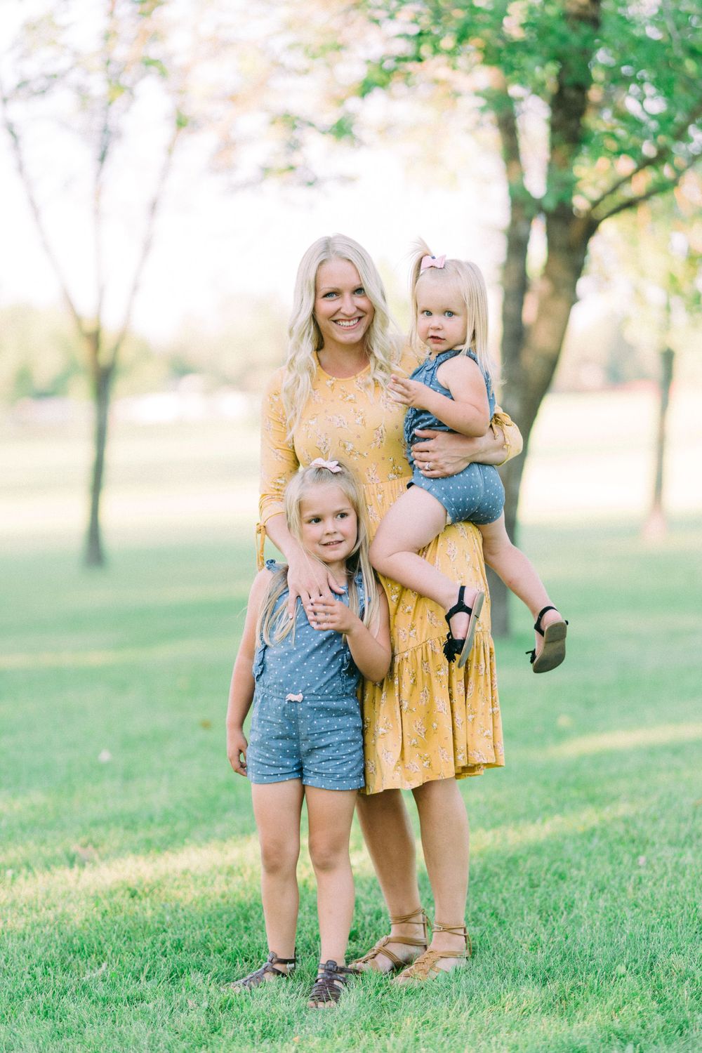 A cheerful mother in a yellow dress poses with her two children wearing denim overalls on a sunny lawn.
