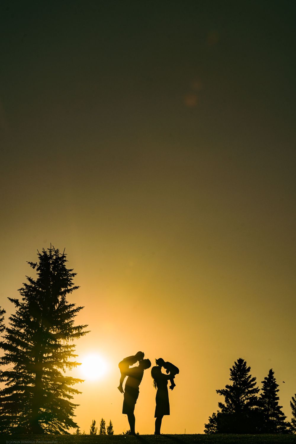 Two silhouettes stand against a golden sunset sky with evergreen trees in the background.