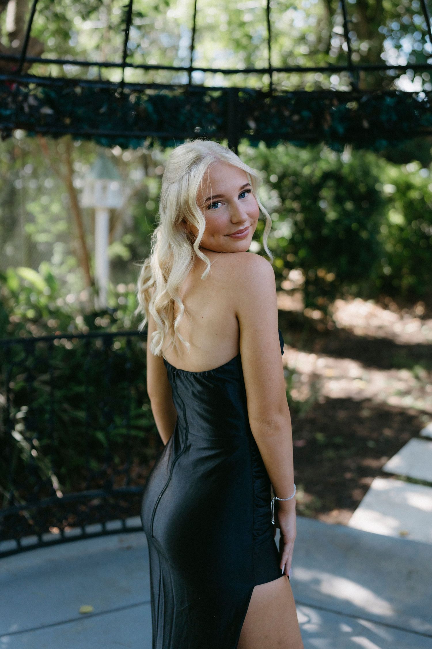 Figure in a fitted black dress poses outdoors against a natural backdrop with sunlit trees and greenery.