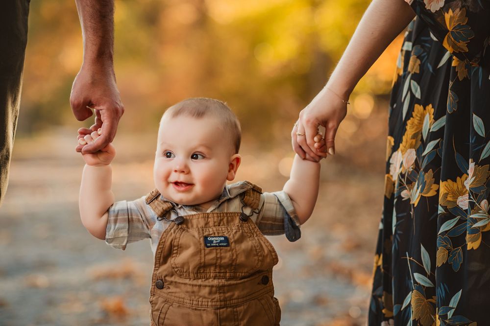 smiling baby boy stands holding his parent's hands for fall minis