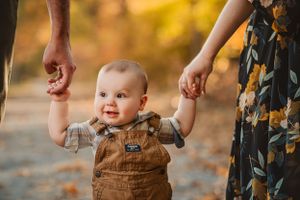 smiling baby boy stands holding his parent's hands for fall minis