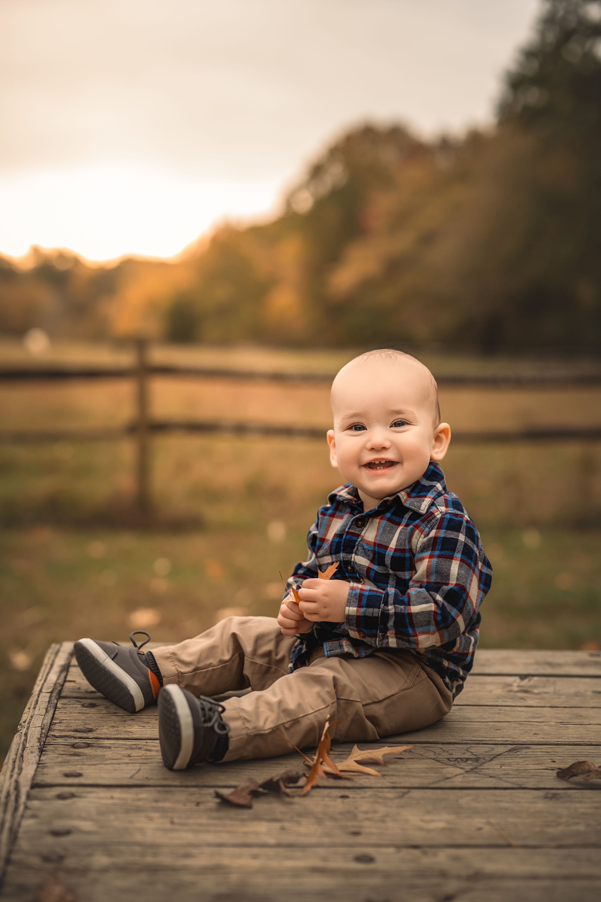 Baby in plaid shirt and khakis lying on wooden deck during sunset photoshoot.