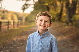 A young boy in a blue gingham shirt smiles brightly outdoors with a wooden fence in the background.