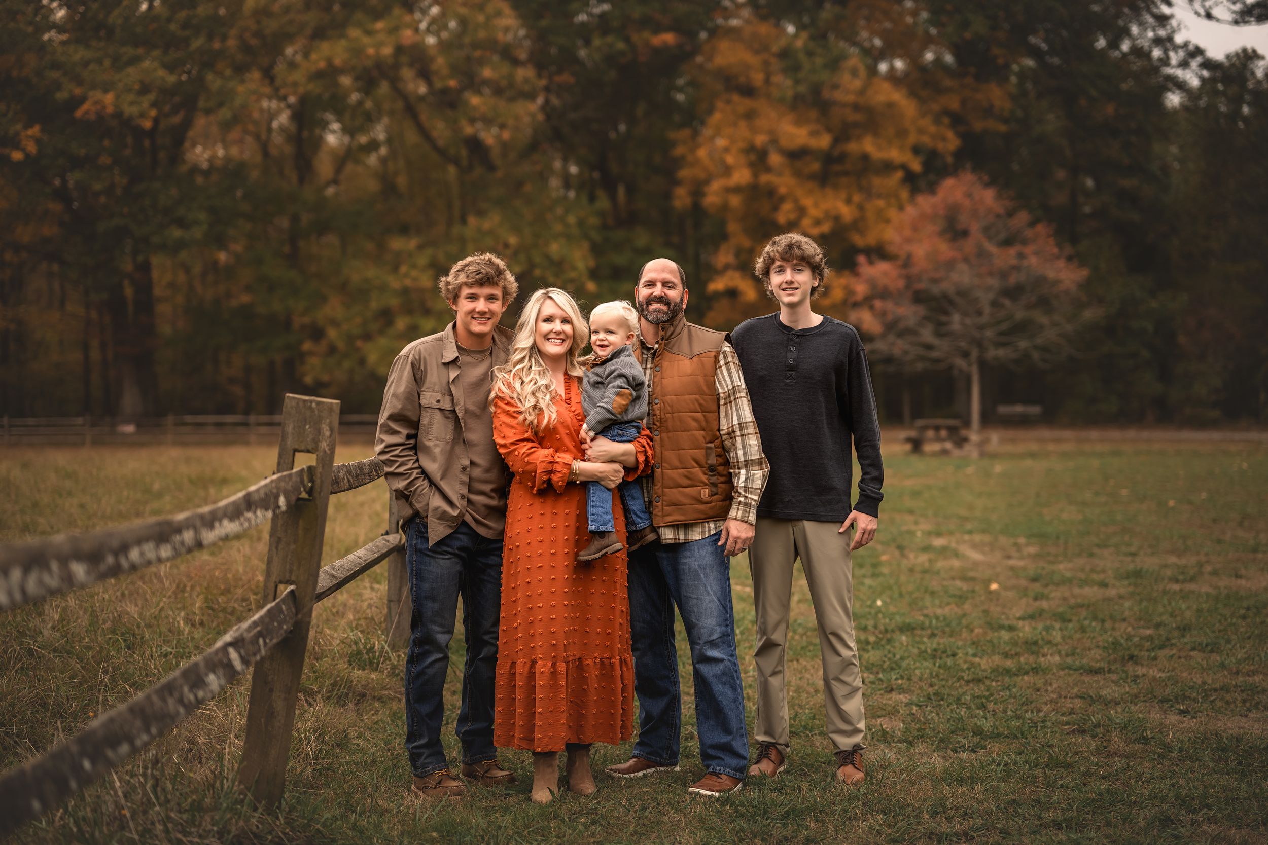 A group wearing coordinated fall colors poses together by a rustic wooden fence during autumn golden hour.