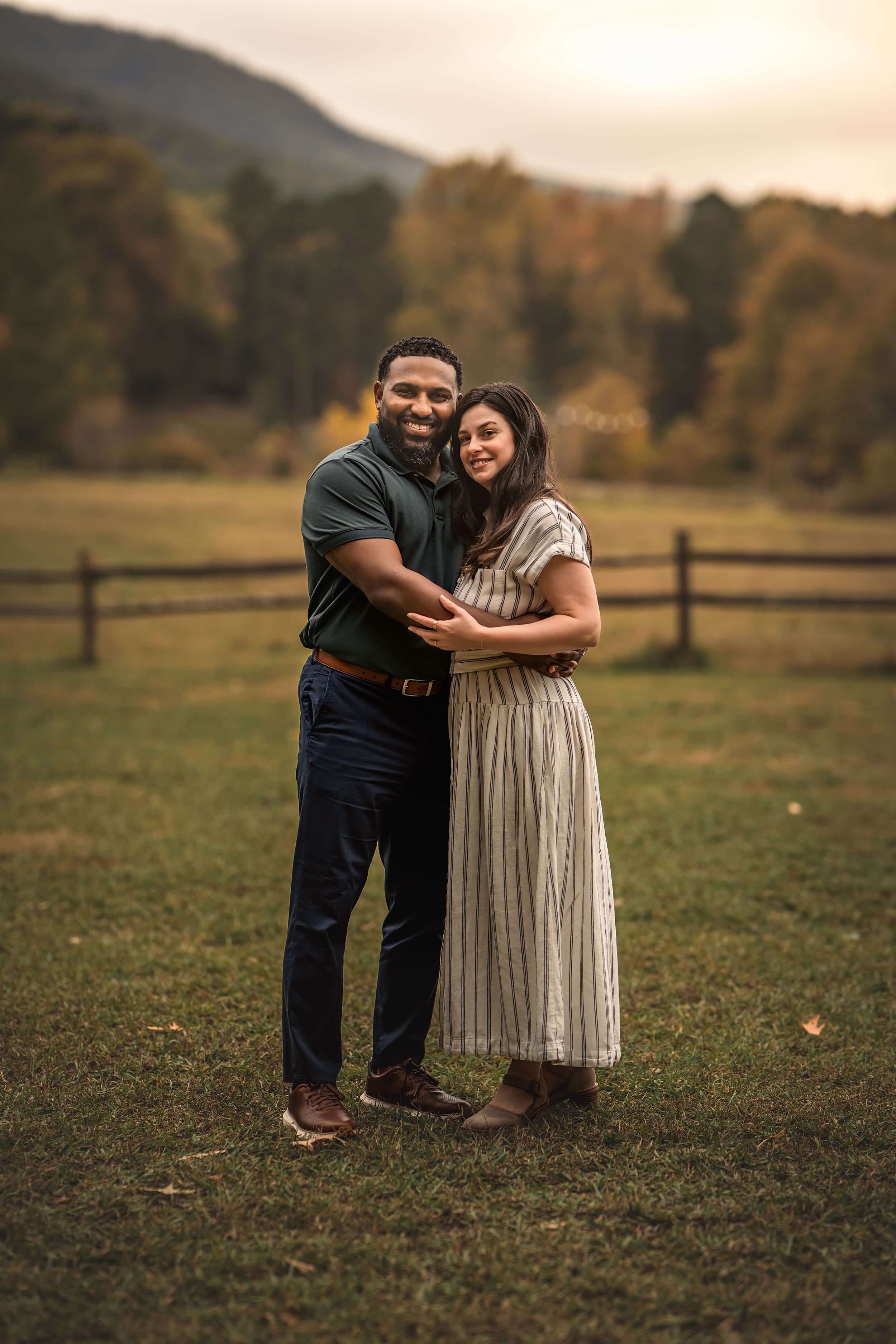 A couple embraces in a gentle pose on grassy autumn fields with wooden fences in the background at sunset.