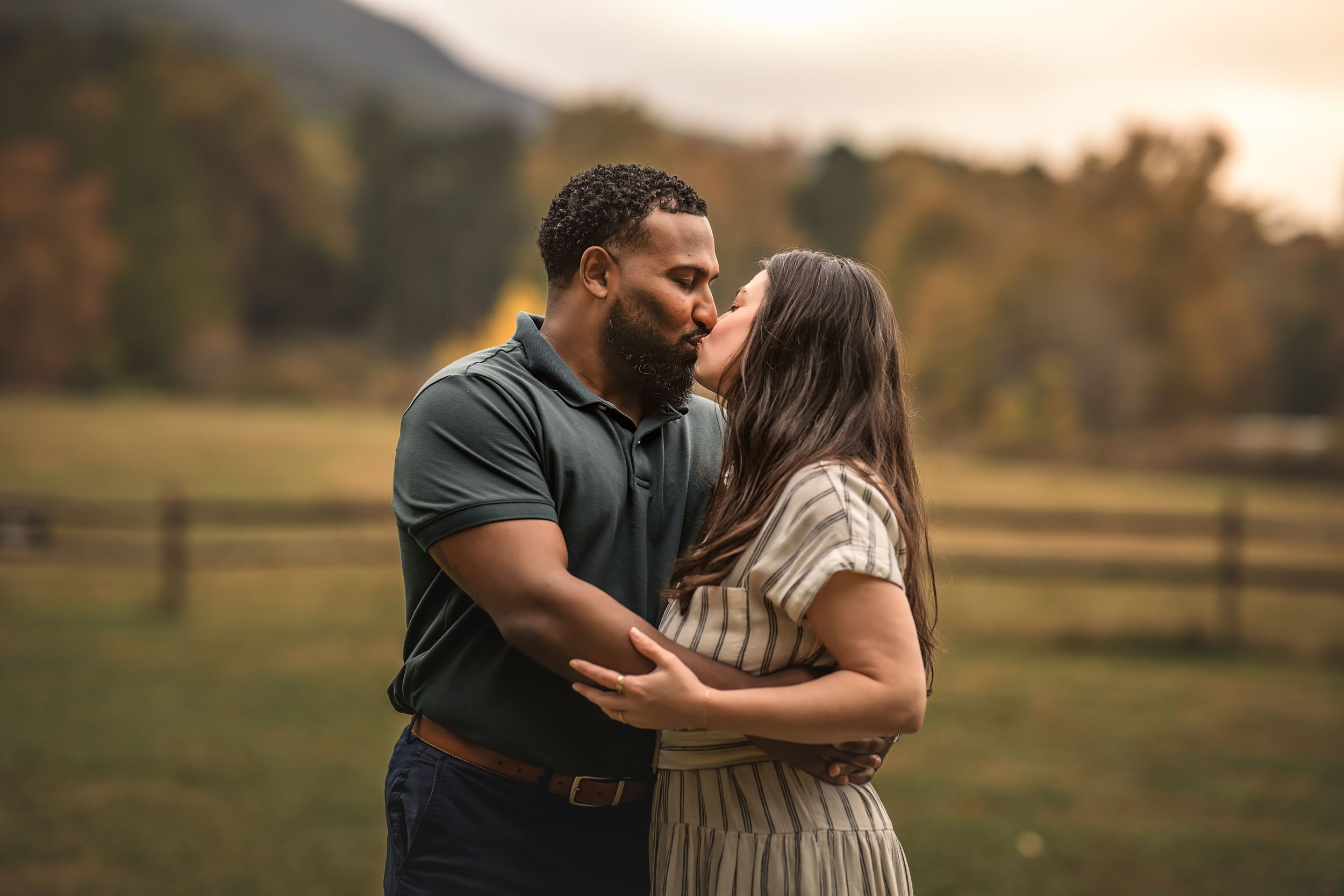 A romantic couple embraces in a rural setting with a wooden fence and autumn hills in the background at sunset.