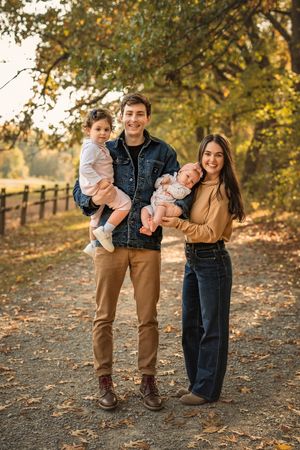 A young family stands together on an autumn path lined with wooden fences and falling leaves at sunset.
