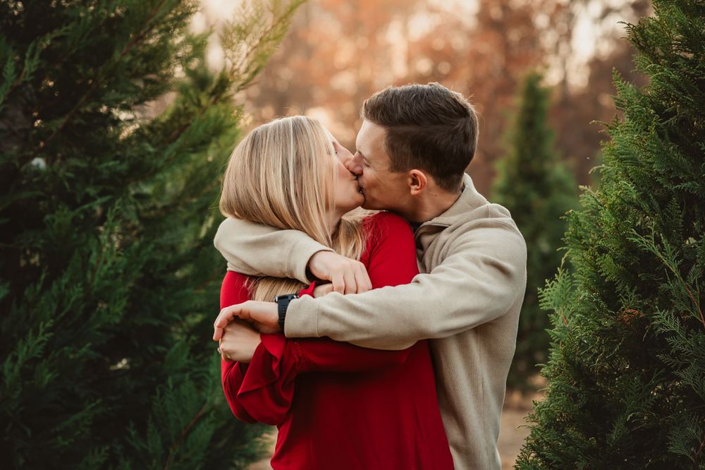 husband hugs wife from behind and kisses her for Christmas tree farm mini session