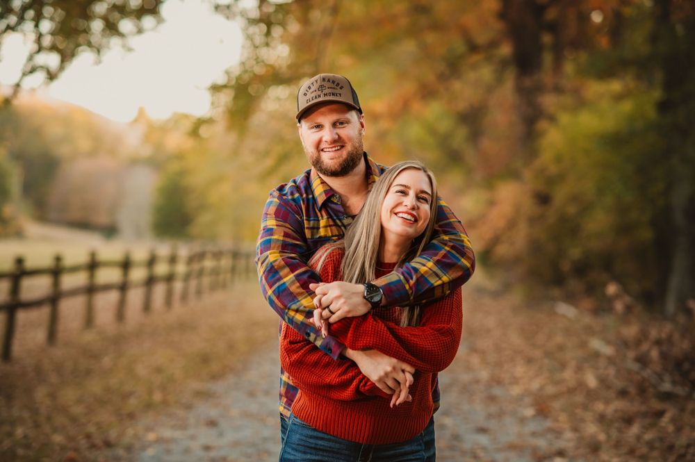 husband hugs wife from behind on tree lined path covered in fall leaves