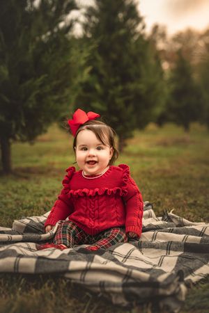 A smiling baby in a red ruffled dress and bow sits on a plaid blanket among evergreen trees.
