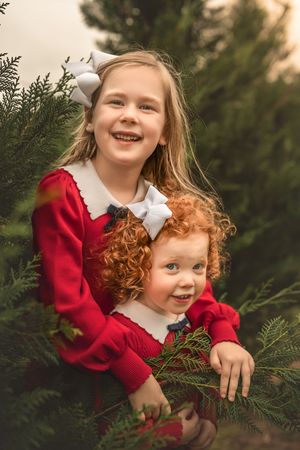 Children wearing red sweaters share a playful moment among evergreen trees during a festive outdoor photo session.