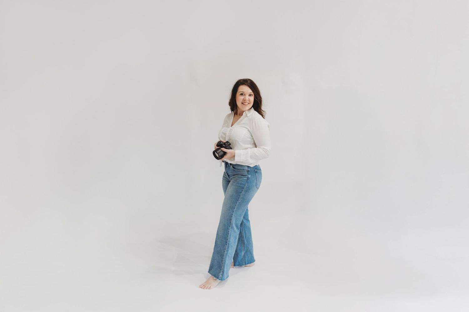Woman in white sweater and jeans poses against plain backdrop in minimalist studio setting. 
Womens headshot | Portrait Photographer