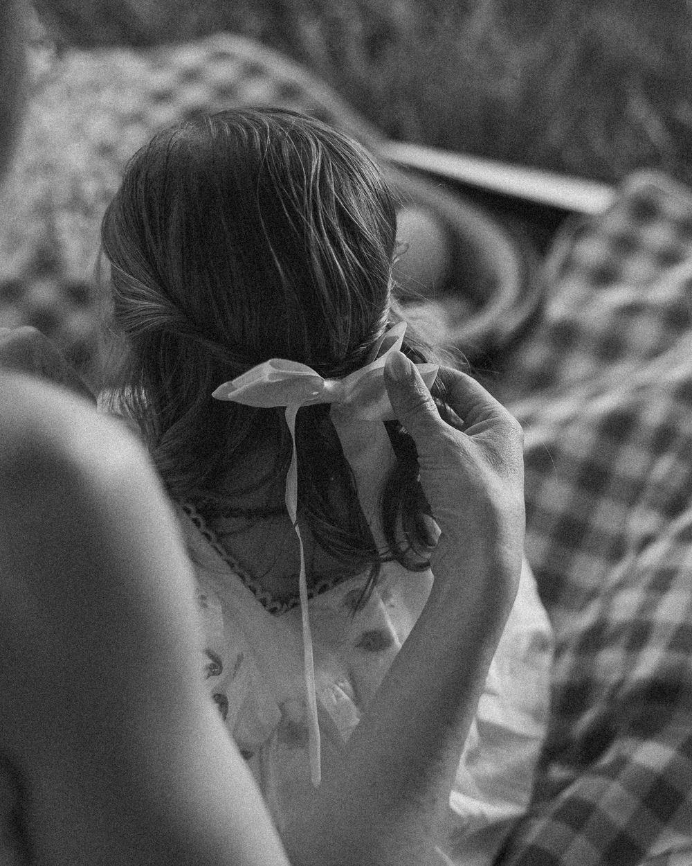 Black and white photo showing a person with a ribbon tied in their hair sitting on a checkered blanket outdoors.