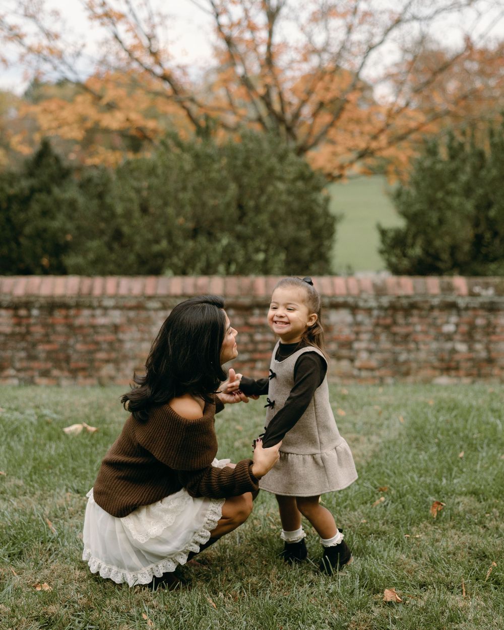 A heartwarming outdoor moment between two people on a grassy lawn with autumn trees and a brick wall in the background.
