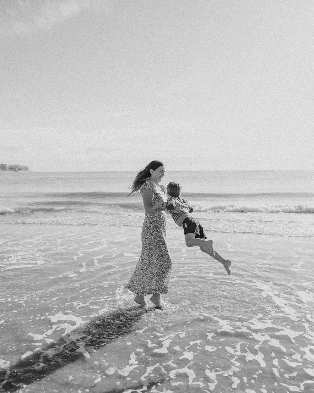 A joyful moment captured in black and white as a woman in a flowing dress swings a child at the beach.