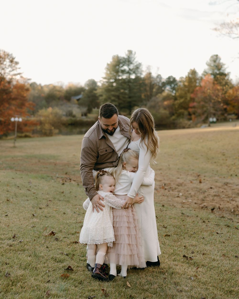 A family of three stands together in a grassy field during autumn wearing coordinating neutral-toned outfits.
